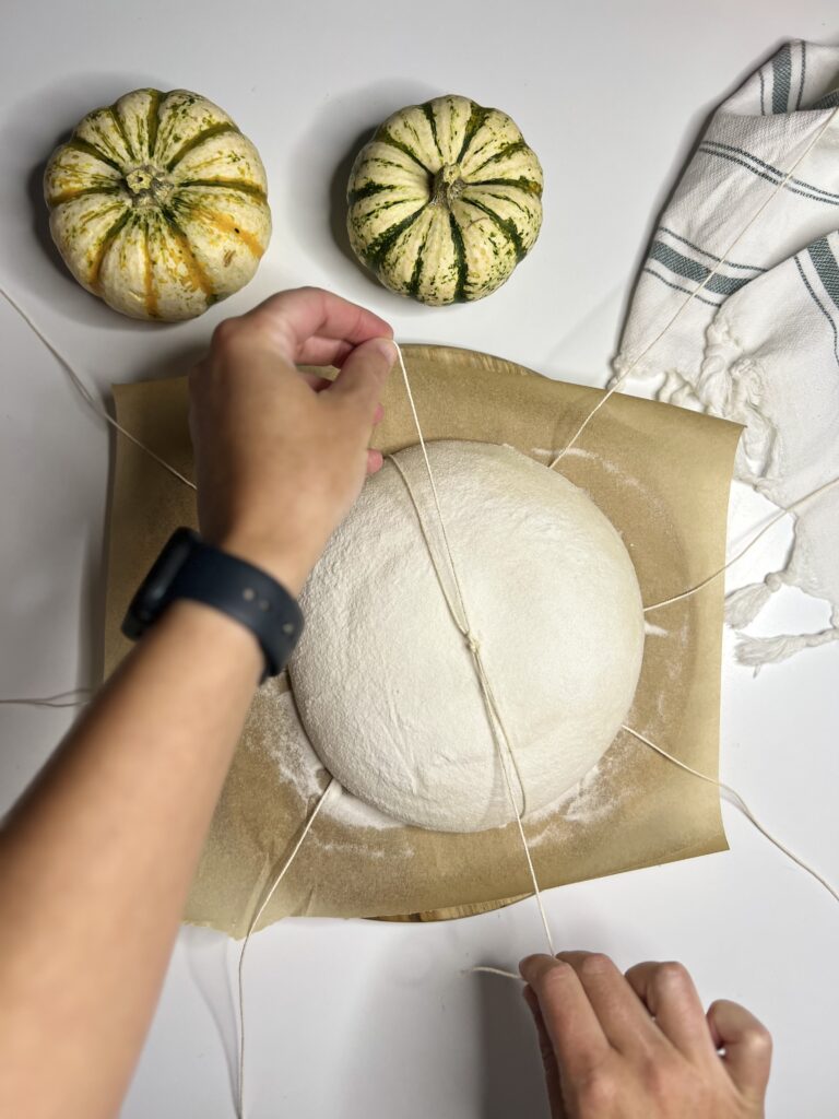 Readjust the strings as needed to ensure the sections are even and begin to tie two at a time. Grab strings from the opposite side of the sourdough boule and tie them in a knot at the center. Do not tie the string too tight as the dough needs room to expand. 